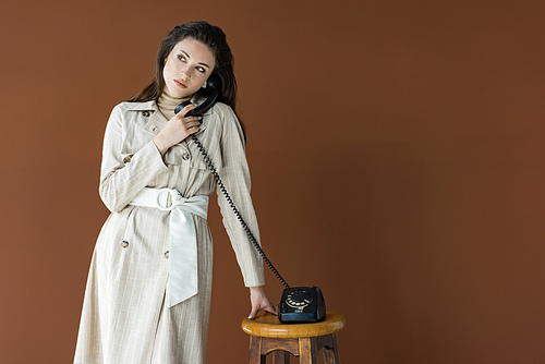 stylish woman in trench coat talking on telephone, looking away and standing isolated on brown