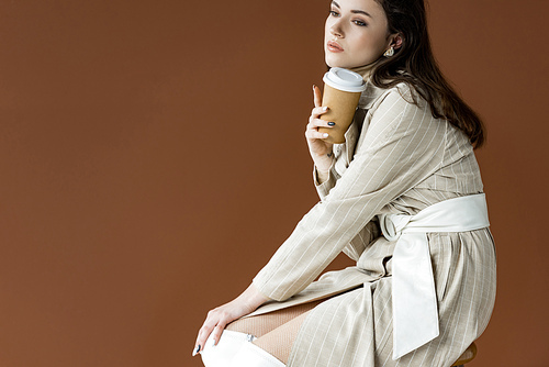 beautiful model looking away, sitting isolated on brown, holding paper cup in hand