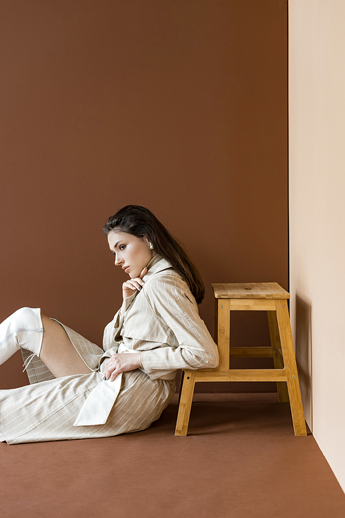 side view of stylish model in beige trench coat sitting on floor, looking away