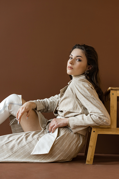 stylish model in trendy trench coat sitting on floor with brown background, 