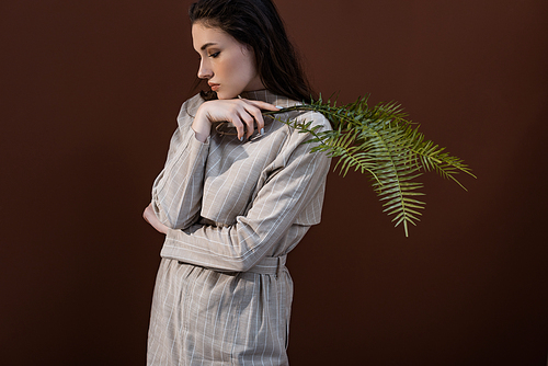 trendy model closing eyes, holding fern leaves in hands, standing on brown background