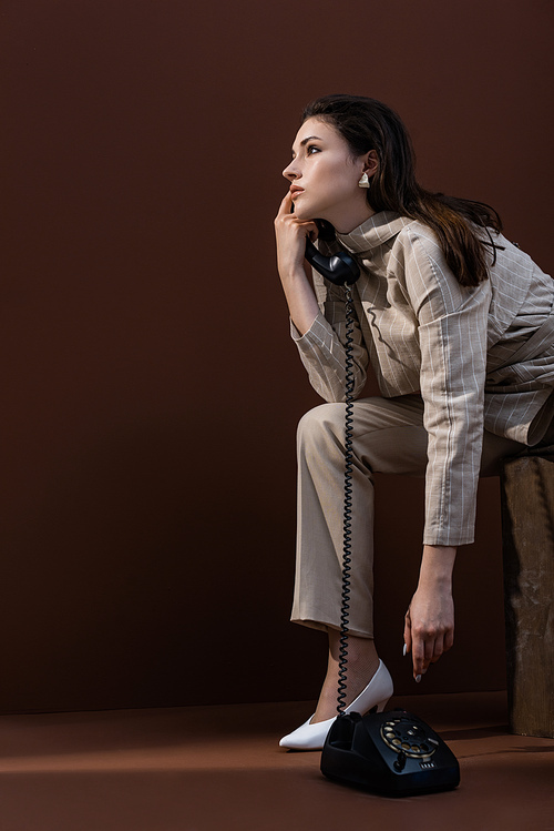 side view of stylish woman in trendy wear talking on telephone, looking away and sitting on brown background