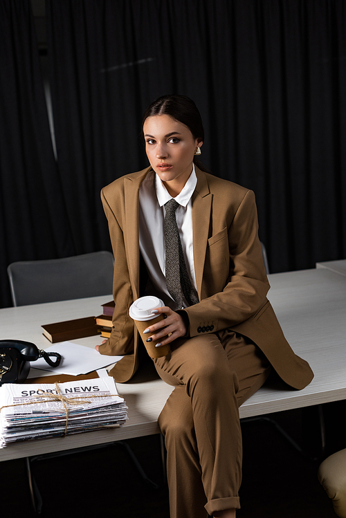 businesswoman in suit with paper cup sitting on table, crossing legs, 