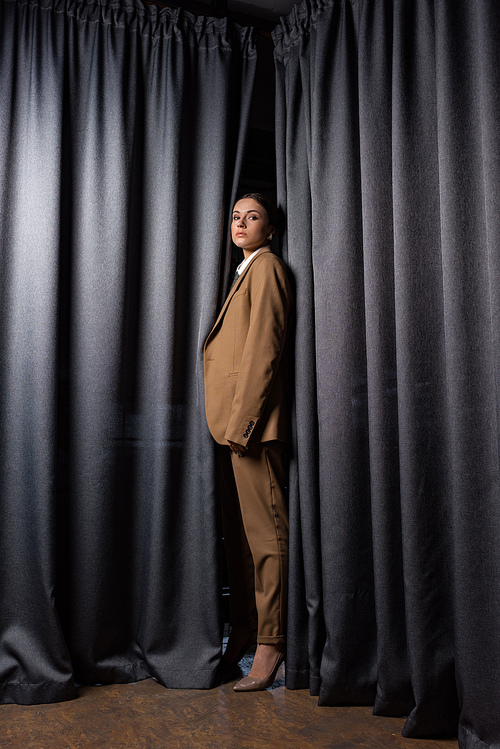 side view of fashionable model in suit standing on dark grey curtain background, 