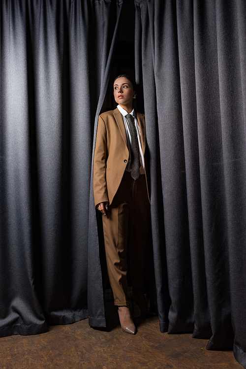 trendy woman in suit standing on curtain background, looking away