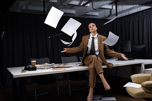 businesswoman sitting on table under falling paper blanks, crossing legs, 