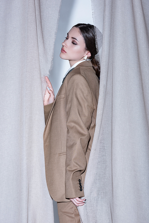 side view of fashionable woman in suit standing on light grey curtain background
