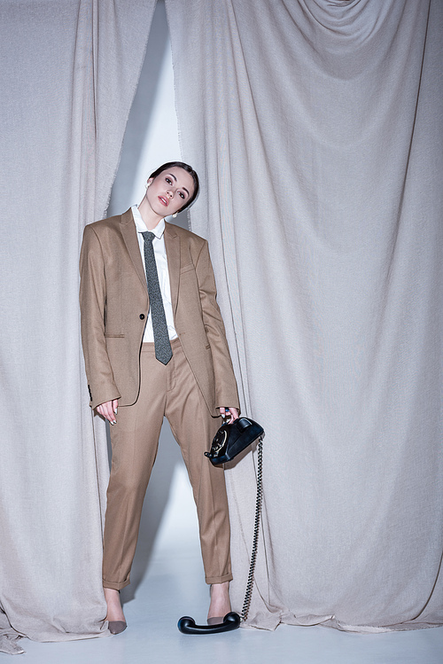 fashionable woman in beige suit standing on curtain background and holding telephone