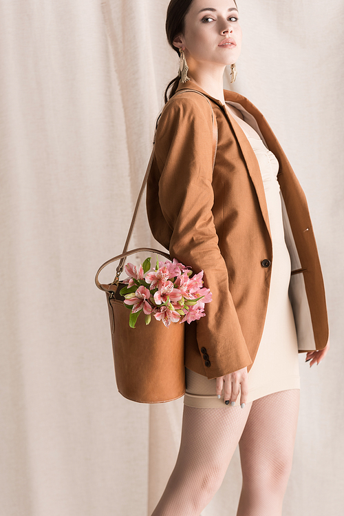 fashionable woman with flowers in bag standing on curtain background, looking away