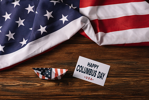 top view of card with happy Columbus Day inscription near paper boat on wooden surface with American national flag