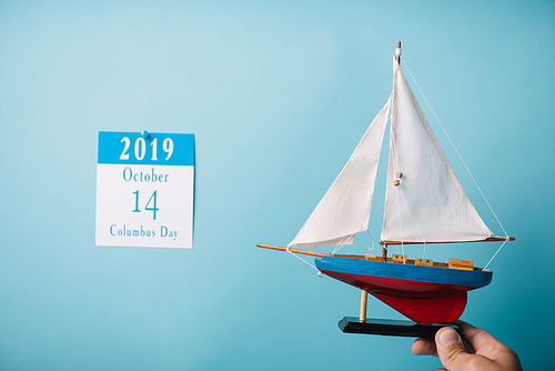 cropped view of man holding crafted ship near paper calendar with Columbus Day inscription isolated on blue