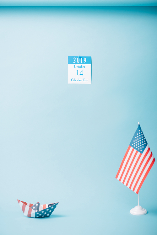 paper calendar with Columbus Day inscription near paper boat and American national flag on blue background