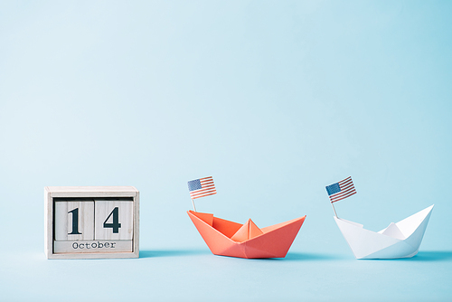 wooden calendar with October 14 date near paper boats with American flag pattern on blue background