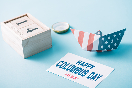 wooden calendar with October 14 date near paper boat with American flag pattern, compass and card with happy Columbus Day inscription on blue background