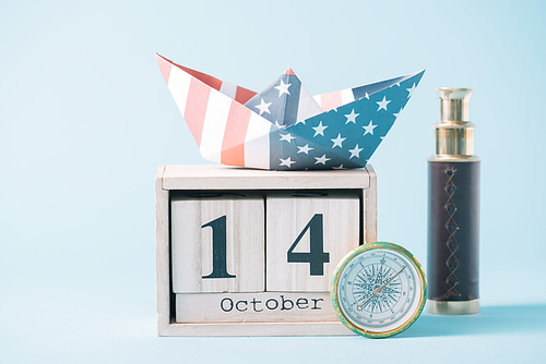 paper boat with American flag pattern on wooden calendar with October 14 date near compass and telescope on blue background