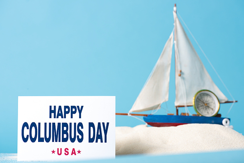 card with happy Columbus Day inscription near miniature ship in white sand isolated on blue