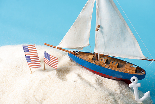 American national flags in white sand near miniature ship and anchor isolated on blue