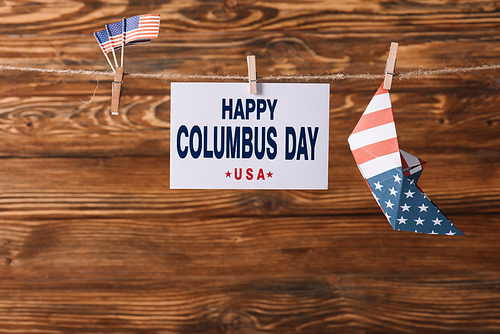 card with happy Columbus day inscription between American national flags and paper boat on wooden surface
