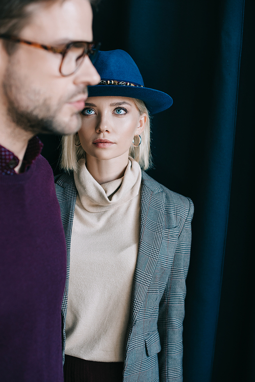 stylish young man in glasses and blonde woman in hat near curtain