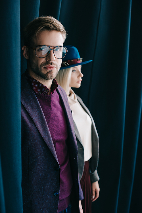 stylish young man in glasses and blonde woman in hat near curtain