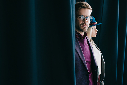 stylish young man in glasses and blonde woman in hat near curtain