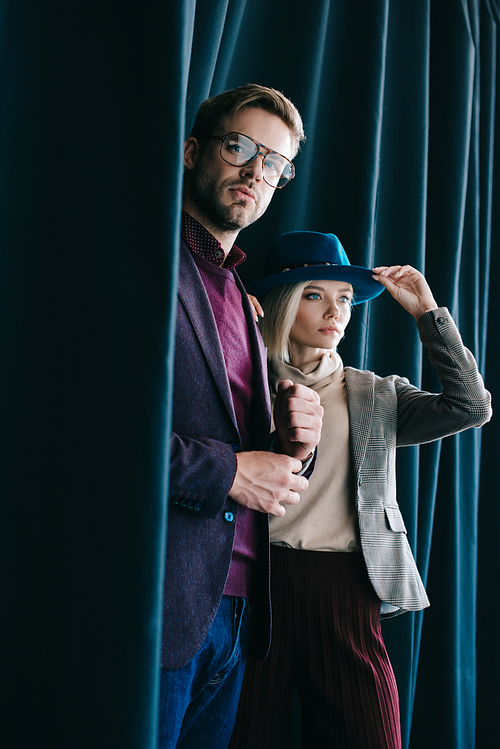 stylish young man in glasses and blonde woman in hat near curtain