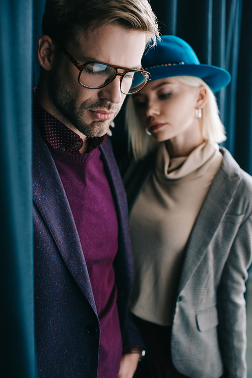 stylish young man in glasses and blonde woman in hat near curtain