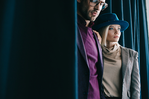 stylish young man in glasses and blonde woman in hat near curtain