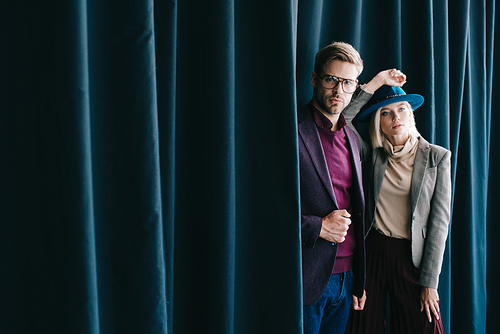 stylish young man in glasses and blonde woman in hat near curtain