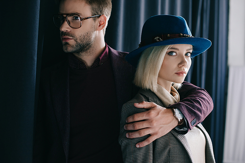 elegant young man in glasses embracing blonde girl in hat near curtain