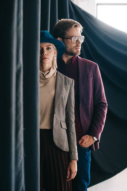 elegant young woman in hat and man in glasses near curtain