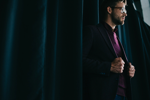 stylish young man in jacket and glasses standing near curtain and looking away