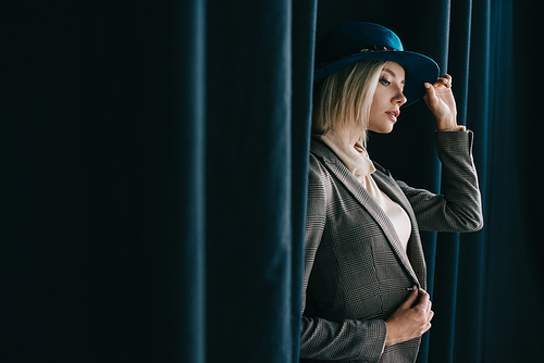 stylish blonde girl in hat and jacket standing near curtain