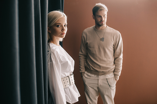 stylish young woman in blouse and man standing near curtain on brown