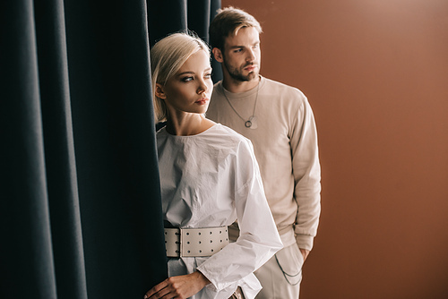 stylish young woman in blouse and man standing near curtain on brown