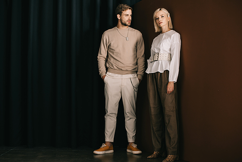 full length view of stylish man with hands in pockets and blonde woman in blouse standing near curtain