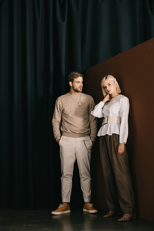 full length view of stylish man with hands in pockets and blonde woman in blouse standing near curtain