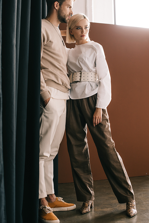 full length view of stylish couple standing near curtain on brown