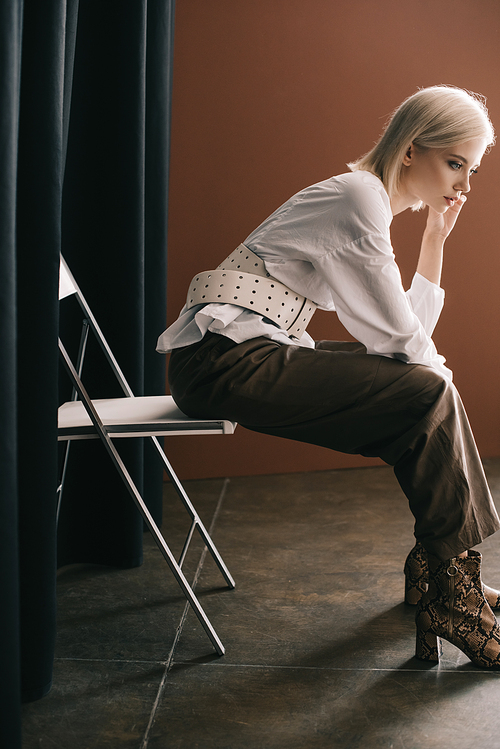 side view of stylish blonde woman in white blouse and boots with snakeskin  sitting on chair near curtain on brown