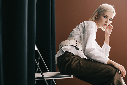 stylish blonde woman in white blouse sitting on chair near curtain on brown