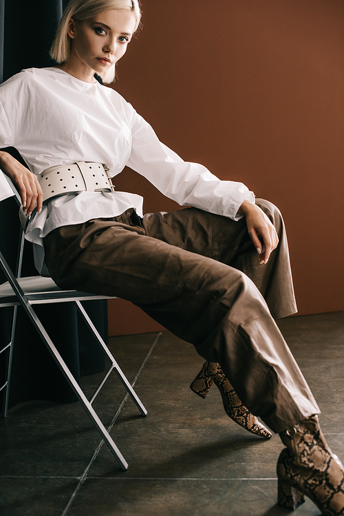 stylish blonde woman in white blouse and boots with snakeskin  sitting on chair near curtain on brown