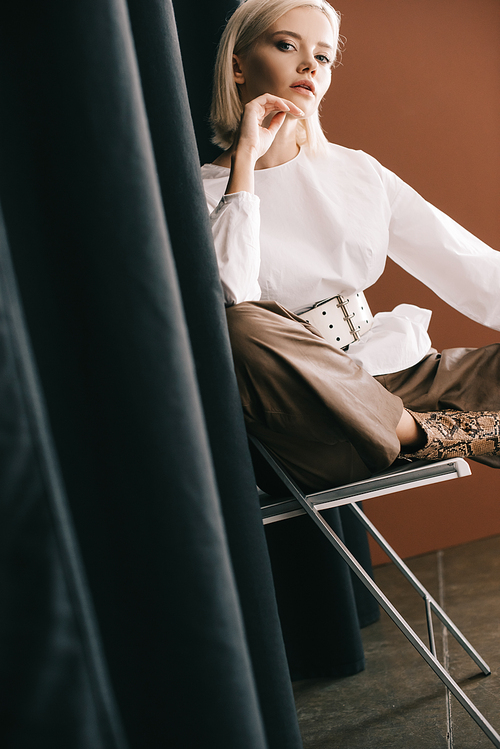 stylish blonde woman in white blouse sitting on chair near curtain on brown