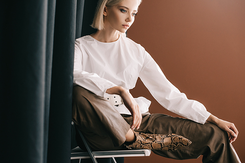 stylish blonde woman in white blouse sitting on chair near curtain on brown