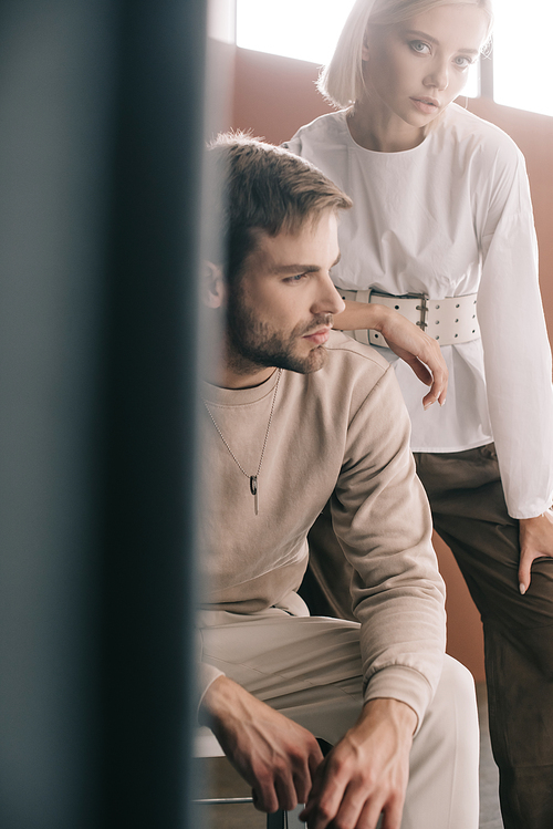 bearded man sitting on chair and attractive blonde woman near curtain on brown