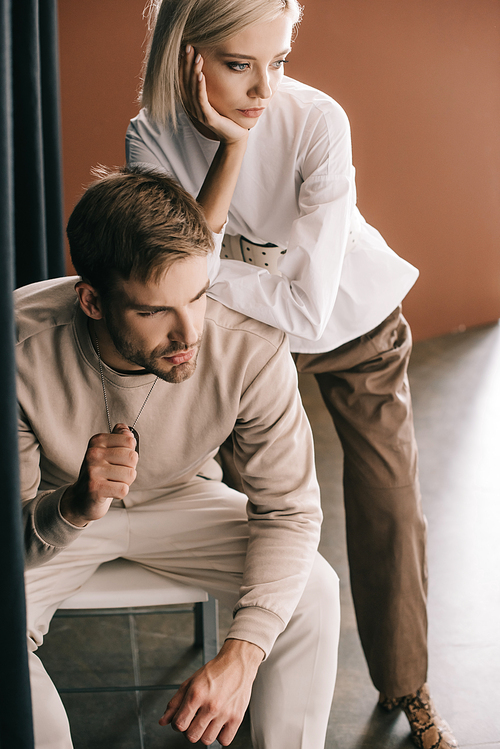 bearded man sitting on chair and attractive blonde woman near curtain on brown