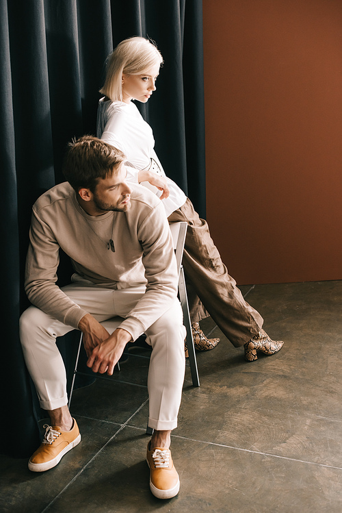 bearded man sitting on chair and attractive blonde woman near curtain on brown