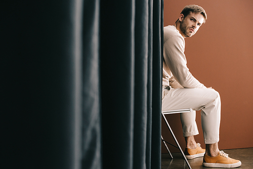 man in casual outfit sitting on chair near curtain on brown