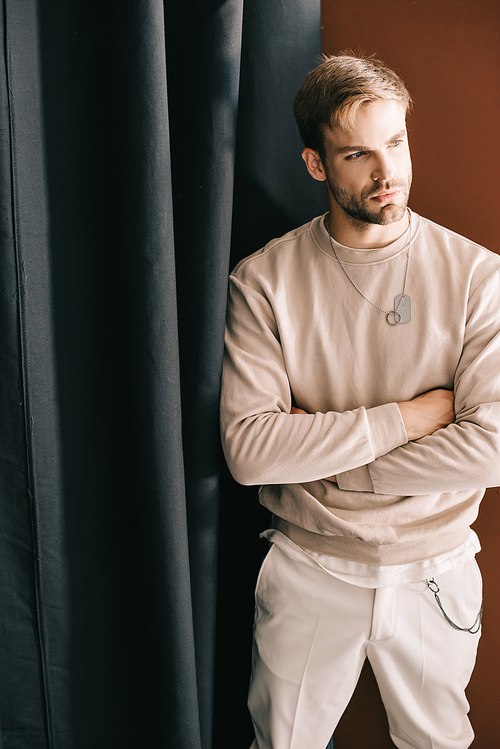 pensive bearded man standing with crossed arms and looking away near curtain