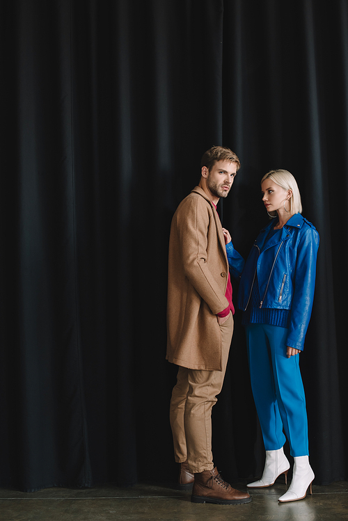 full length view of stylish young woman and man in autumn outfits near black curtain