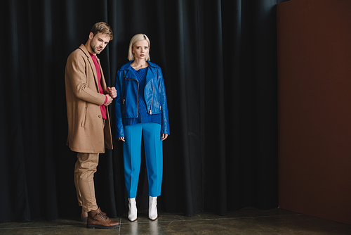 full length view of stylish young woman and man in autumn outfits near black curtain